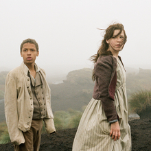 Two children in historical clothing stand on a misty, rocky landscape with sparse grass and dark soil.