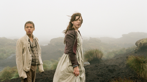 Two children in historical clothing stand on a misty, rocky landscape with sparse grass and dark soil.
