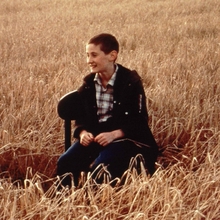 A boy sitting on a chair in the middle of a golden wheat field, wearing a plaid shirt and dark jacket.