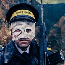 A traffic warden in uniform and cap stands amid a war-torn street with smoke, debris, and flames in the background.