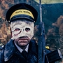 A traffic warden in uniform and cap stands amid a war-torn street with smoke, debris, and flames in the background.