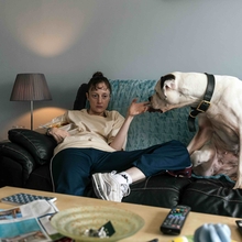 Woman sitting on a couch holding her hand by a large white dog, with a lamp, TV remotes, and newspapers on a nearby table.