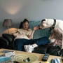 Woman sitting on a couch holding her hand by a large white dog, with a lamp, TV remotes, and newspapers on a nearby table.