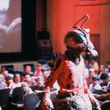 A still from Matinee of a person in detailed red insect costume standing in a cinema with an audience watching a black-and-white film.