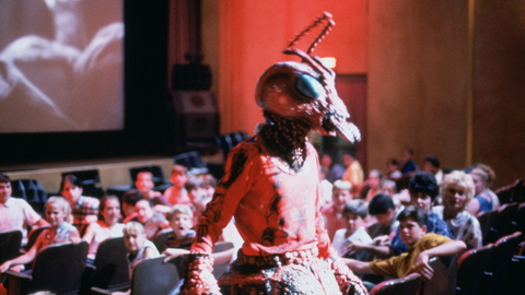 A still from Matinee of a person in detailed red insect costume standing in a cinema with an audience watching a black-and-white film.