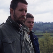 Two people in jackets standing outdoors with a blurred countryside landscape in the background under a cloudy sky.