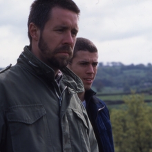 Two people in jackets standing outdoors with a blurred countryside landscape in the background under a cloudy sky.