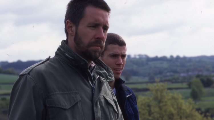 Two people in jackets standing outdoors with a blurred countryside landscape in the background under a cloudy sky.