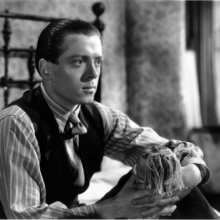 Black and white photo of a man in a vest and striped shirt sitting on a bed, holding a doll with yarn hair in his hands.