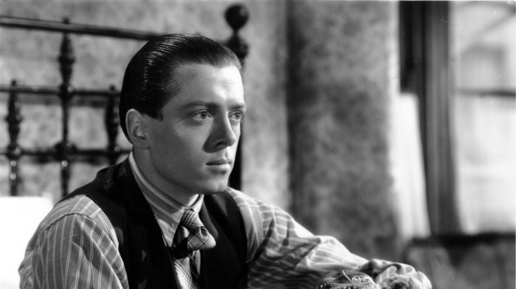 Black and white photo of a man in a vest and striped shirt sitting on a bed, holding a doll with yarn hair in his hands.