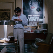A still from Closed Circuit of two men in a cluttered office, one sitting and one standing, surrounded by film reels, a vintage lamp, and posters on the wall.