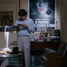 A still from Closed Circuit of two men in a cluttered office, one sitting and one standing, surrounded by film reels, a vintage lamp, and posters on the wall.