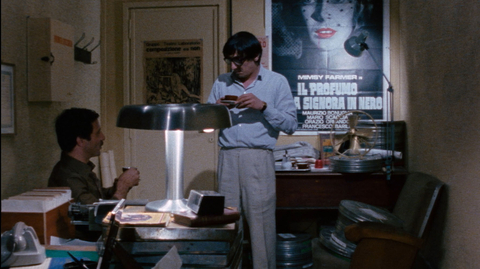 A still from Closed Circuit of two men in a cluttered office, one sitting and one standing, surrounded by film reels, a vintage lamp, and posters on the wall.