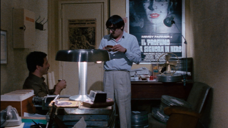 A still from Closed Circuit of two men in a cluttered office, one sitting and one standing, surrounded by film reels, a vintage lamp, and posters on the wall.