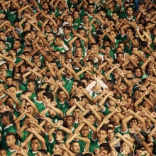 Crowd of sports fans wearing green jerseys, raising their arms and crossing them in unison, showing unified support.