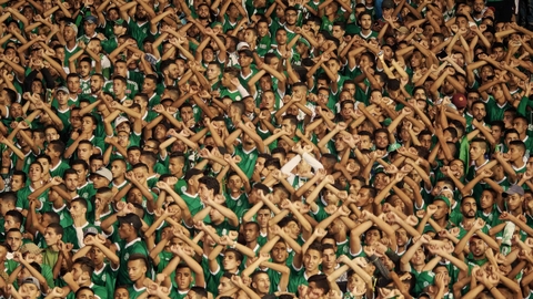 Crowd of sports fans wearing green jerseys, raising their arms and crossing them in unison, showing unified support.