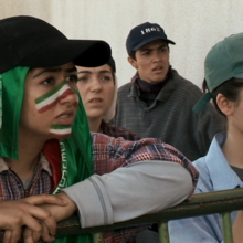 A group of four people leaning on a green metal railing, wearing casual clothes and baseball caps.