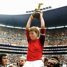 Footballer in red jersey raised on shoulders holding a trophy in a crowded stadium celebrating a sports victory.