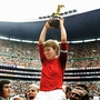 Footballer in red jersey raised on shoulders holding a trophy in a crowded stadium celebrating a sports victory.