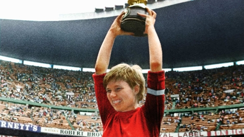 Footballer in red jersey raised on shoulders holding a trophy in a crowded stadium celebrating a sports victory.