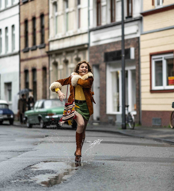 Person joyfully splashing through a puddle on a street lined with parked vintage cars and old buildings.