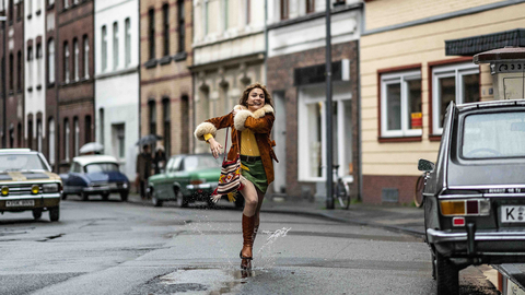 Person joyfully splashing through a puddle on a street lined with parked vintage cars and old buildings.