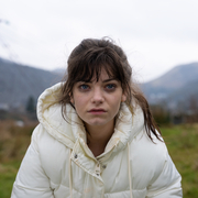 A still from Effi O Blaenau of a young woman wearing a white puffer jacket in a grassy outdoor area with mountains and power lines in the background on a cloudy day.