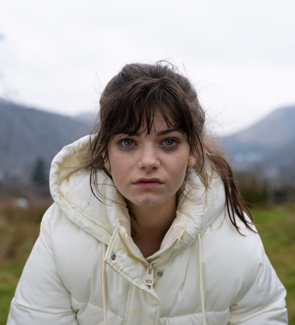 A still from Effi O Blaenau of a young woman wearing a white puffer jacket in a grassy outdoor area with mountains and power lines in the background on a cloudy day.