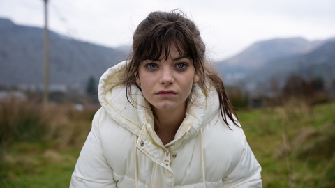 A still from Effi O Blaenau of a young woman wearing a white puffer jacket in a grassy outdoor area with mountains and power lines in the background on a cloudy day.