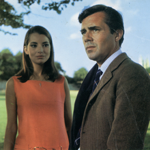 Two people standing outdoors on a sunny day with trees and grass in the background, one in an orange dress and the other in a brown suit.