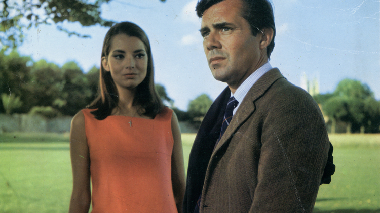 Two people standing outdoors on a sunny day with trees and grass in the background, one in an orange dress and the other in a brown suit.