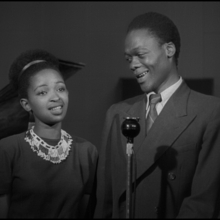 Black and white photo of a woman and man standing side by side, with a microphone in front and a piano in the background.