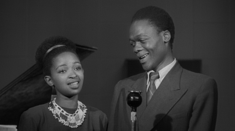 Black and white photo of a woman and man standing side by side, with a microphone in front and a piano in the background.