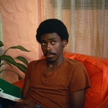 Person in a brown shirt sits on an orange couch reading a magazine, with a plant and a textured wall in the background.