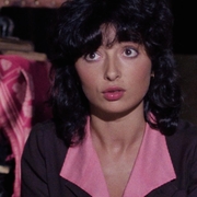 A still from Closed Circuit of a woman with dark curly hair wearing a brown jacket and pink collar shirt sitting indoors near a table with a patterned tablecloth.
