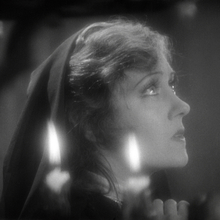 Black and white close-up of a person with long hair partially covered by a dark hood, dimly lit by a candle flame.