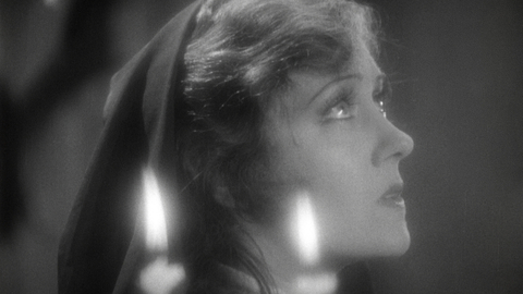 Black and white close-up of a person with long hair partially covered by a dark hood, dimly lit by a candle flame.