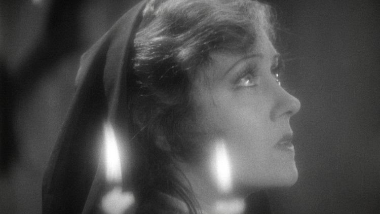 Black and white close-up of a person with long hair partially covered by a dark hood, dimly lit by a candle flame.