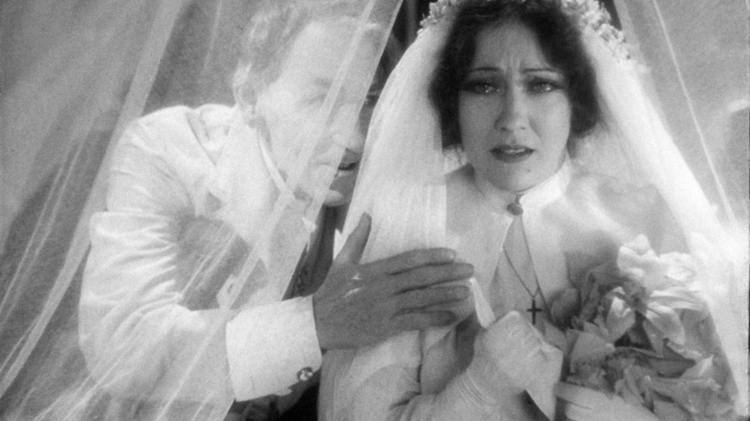 Black and white photo of a bride and groom under a veil, the bride holding a bouquet and wearing a cross necklace.