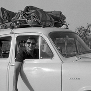 Black-and-white photo of young men traveling in a vintage compact car with luggage strapped to the roof, roadside rural setting.