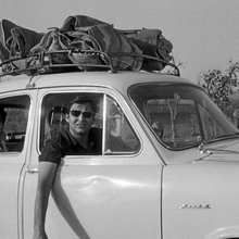 Black-and-white photo of young men traveling in a vintage compact car with luggage strapped to the roof, roadside rural setting.