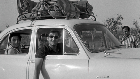 Black-and-white photo of young men traveling in a vintage compact car with luggage strapped to the roof, roadside rural setting.