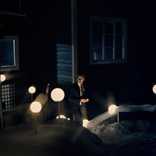 Man in a dark suit standing outside a snow-covered building at night, illuminated by glowing round pathway lights, cinematic noir atmosphere with dramatic low-light photography.