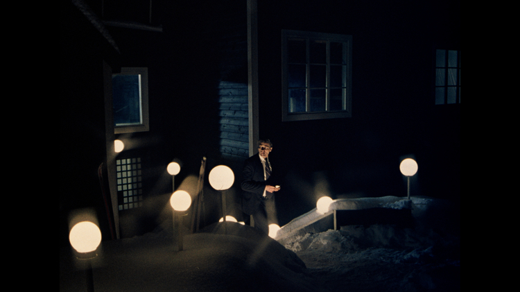 Man in a dark suit standing outside a snow-covered building at night, illuminated by glowing round pathway lights, cinematic noir atmosphere with dramatic low-light photography.