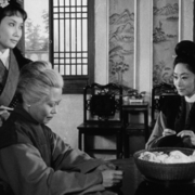Black-and-white photo of three people in traditional East Asian clothing inside a room with wooden chairs and lattice windows.