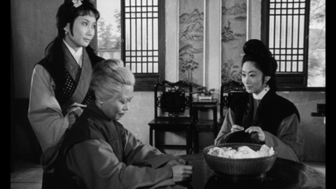 Black-and-white photo of three people in traditional East Asian clothing inside a room with wooden chairs and lattice windows.