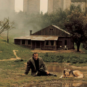 Person sitting on grass beside a dog near a small wooden house, with misty trees and tall stone structures in the background.