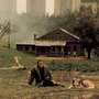 Person sitting on grass beside a dog near a small wooden house, with misty trees and tall stone structures in the background.