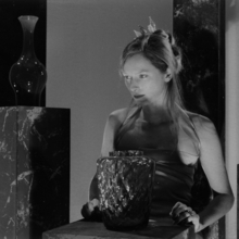 Black and white photo of a woman in a sleeveless dress sitting behind a table with a large textured glass vase.