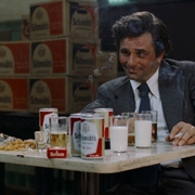 Two men sit at a table with cans of Schmidt's beer, glasses of milk and beer, a cigarette, and snacks in a dimly lit room.